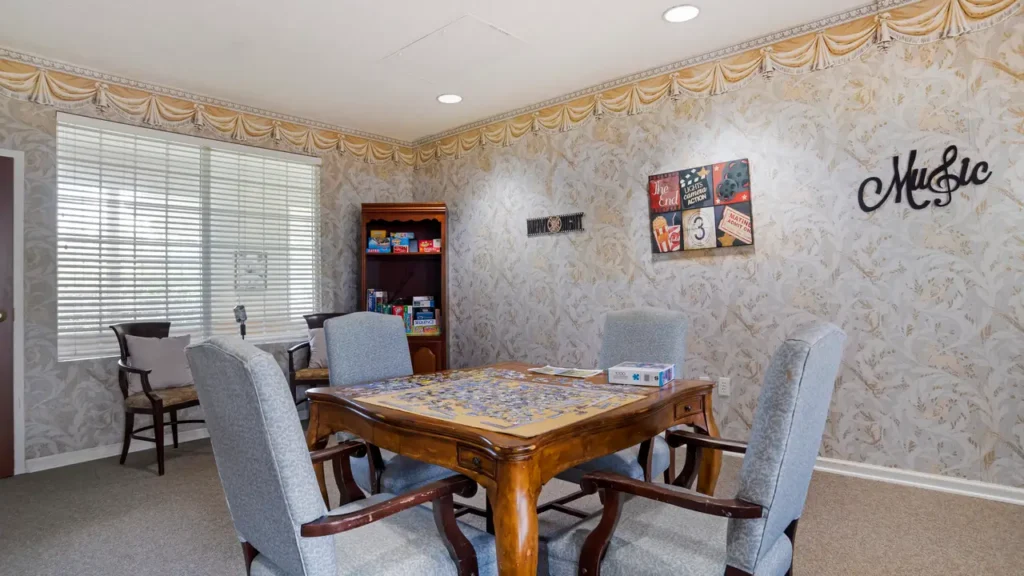 A cozy game room with four gray upholstered chairs around a wooden table with a puzzle in progress. Board games fill a bookshelf in the corner, and music-themed wall art decorates the wallpapered walls.