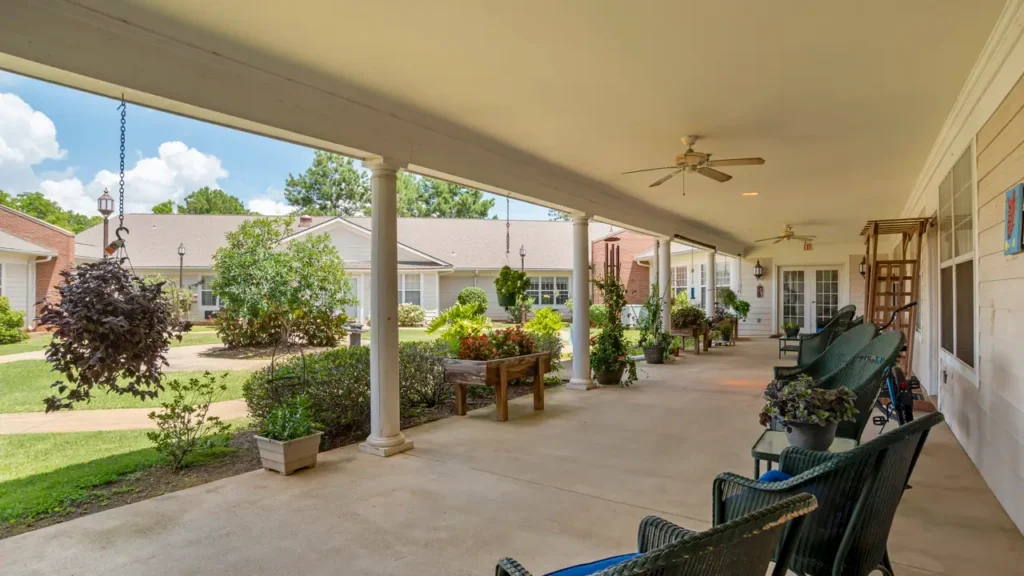A covered patio with ceiling fans, wicker chairs, and potted plants overlooks a landscaped courtyard with bushes and small trees on a sunny day.