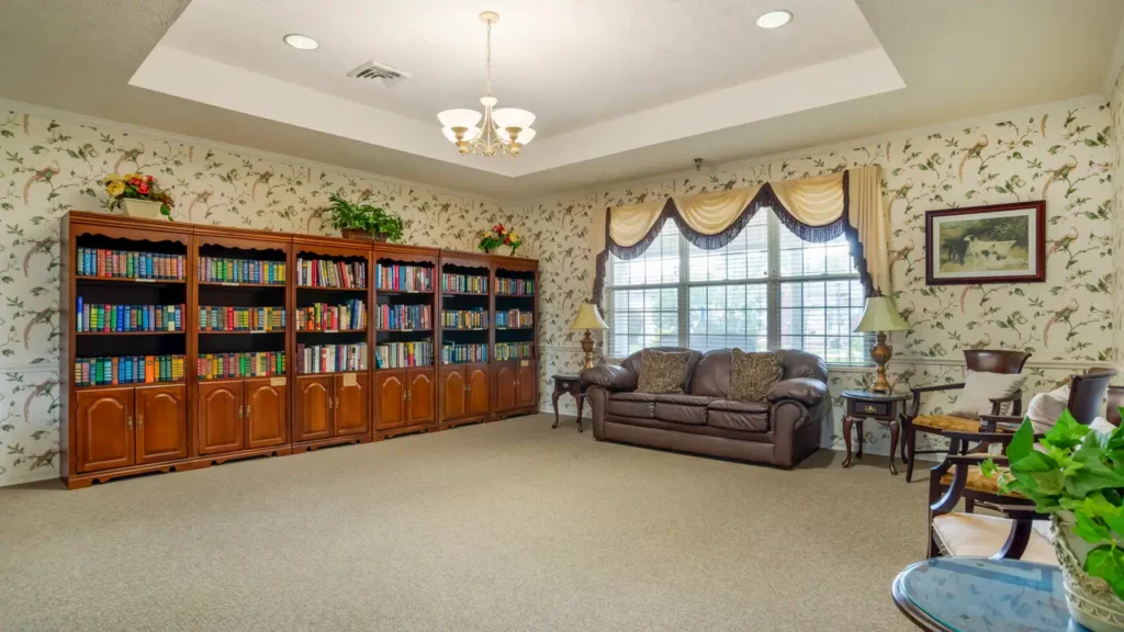 A cozy room with a large bookshelf filled with colorful books, a brown leather sofa with cushions, two side tables with lamps, and a big window with drapes, set against floral wallpaper.