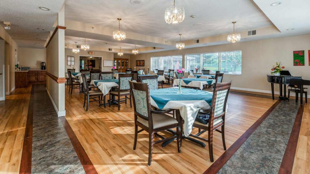 A bright, spacious dining room with wooden floors, tables covered in teal cloths, chairs arranged neatly, large windows letting in natural light, and elegant chandeliers hanging from the ceiling.
