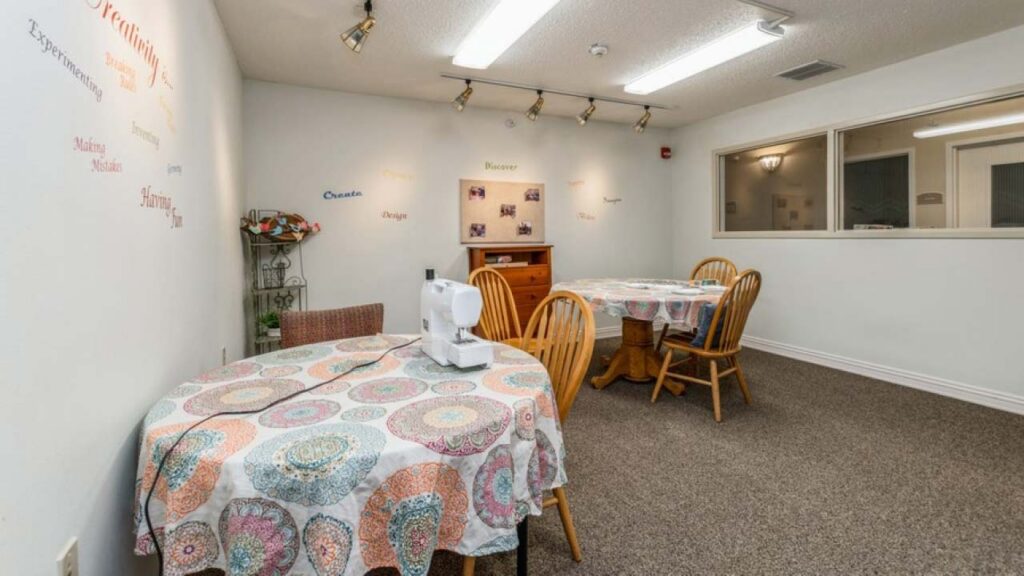 A craft room with two round tables covered in patterned cloths, a sewing machine on one table, wooden chairs, beige carpet, wall art, a small cabinet, and track lights on the ceiling.