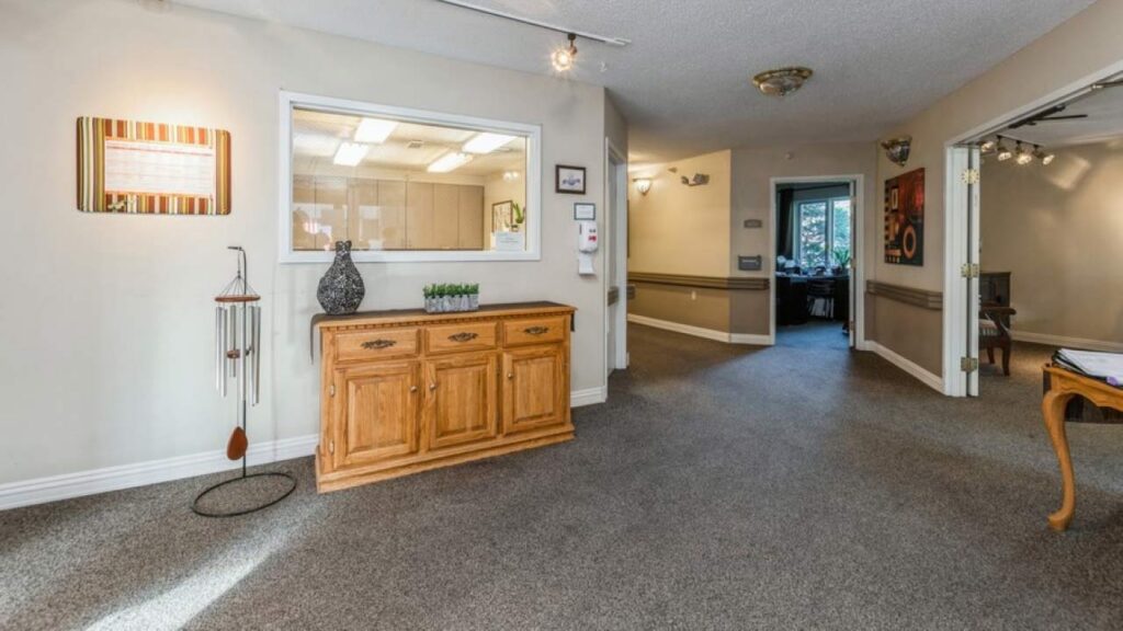 A spacious, carpeted hallway features a wooden sideboard with decorative items, a wall-mounted box, a window to an office, and open doorways leading to other rooms in a well-lit interior space.