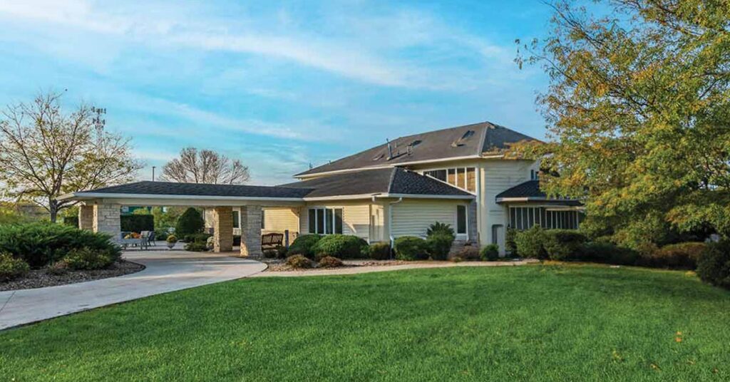 A large, modern house with beige siding, a dark roof, an attached covered driveway, and well-maintained landscaping surrounded by green grass and trees under a blue sky.