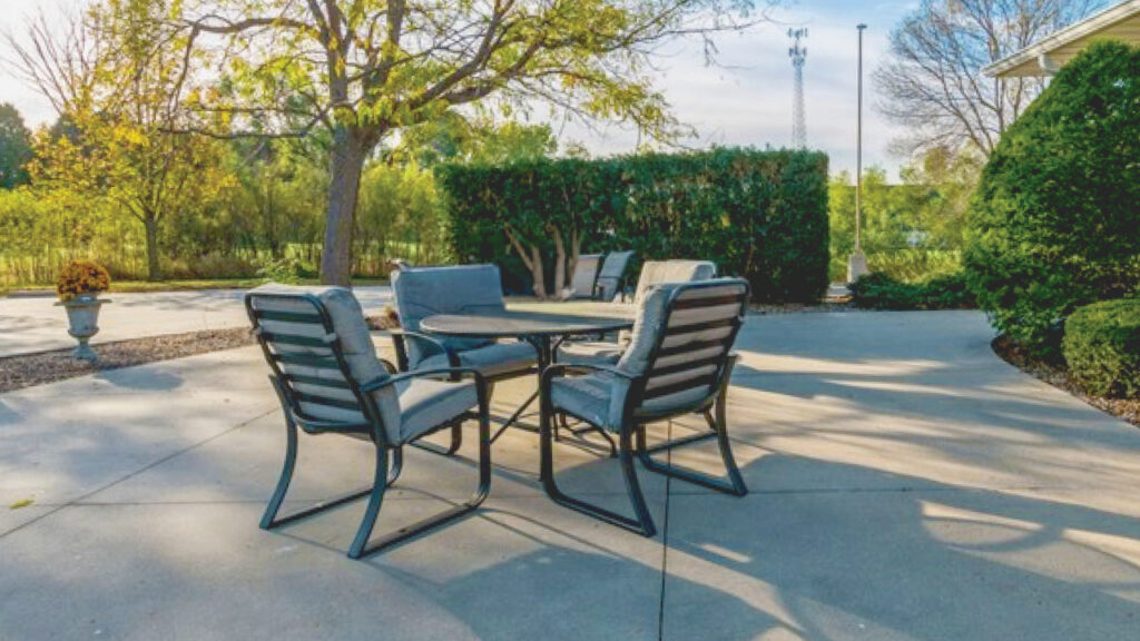 A patio with a round table and four cushioned chairs sits on a concrete surface. Trees, bushes, and greenery surround the area, with sunlight casting shadows across the patio.