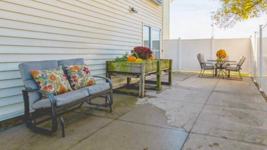 A concrete patio with a cushioned bench, a planter box with pumpkins and flowers, and an outdoor dining table with chairs and a vase of yellow flowers, next to a white fence and house.