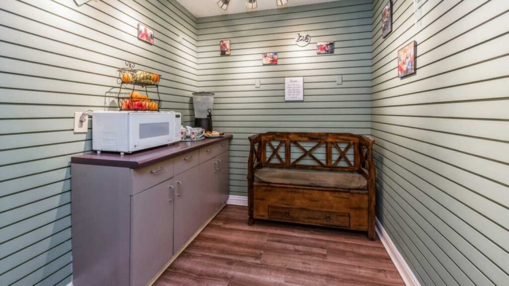 A small room with mint green slatted walls, a brown wooden bench, a countertop with a microwave, water dispenser, and fruit stand, and family photos hanging on the walls. The floor is wood laminate.