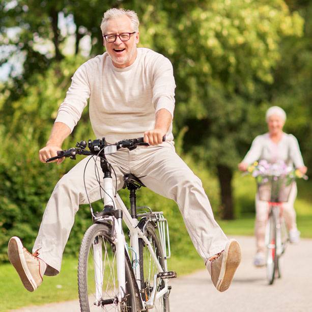An older man rides a bicycle with his legs joyfully outstretched, smiling, while an older woman cycles behind him on a park path surrounded by greenery.