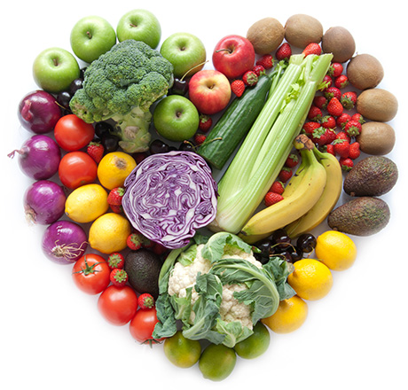 A variety of fresh fruits and vegetables arranged in the shape of a heart, including apples, broccoli, strawberries, tomatoes, bananas, lemons, cauliflower, avocados, cucumbers, kiwi, and purple cabbage.