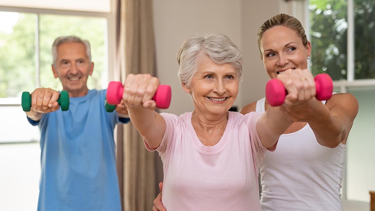 Three smiling adults exercise indoors, holding small dumbbells with arms extended forward. An older woman in the center is assisted by a younger woman, while an older man stands beside them. Sunlight streams through windows in the background.
