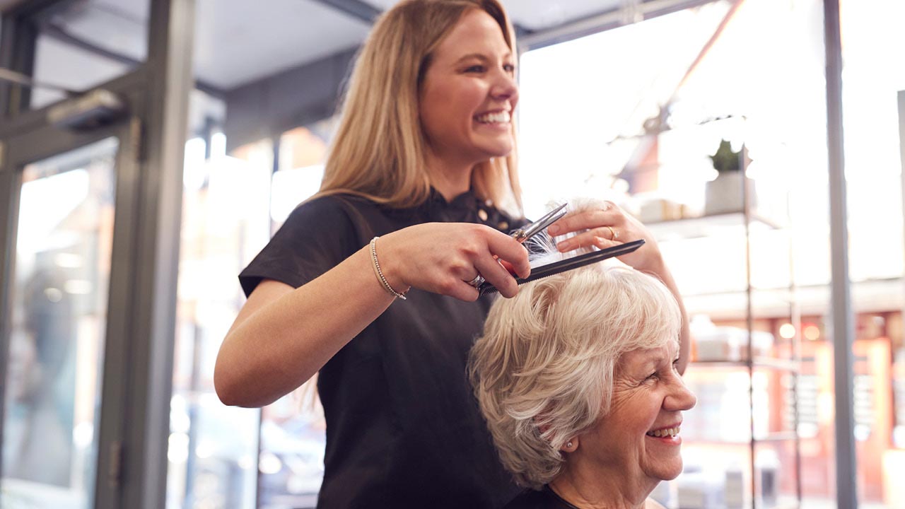 A smiling hairdresser trims the hair of an older woman in a salon. Sunlight streams through large windows, and shelves with decor are visible in the background. Both women appear happy and relaxed.