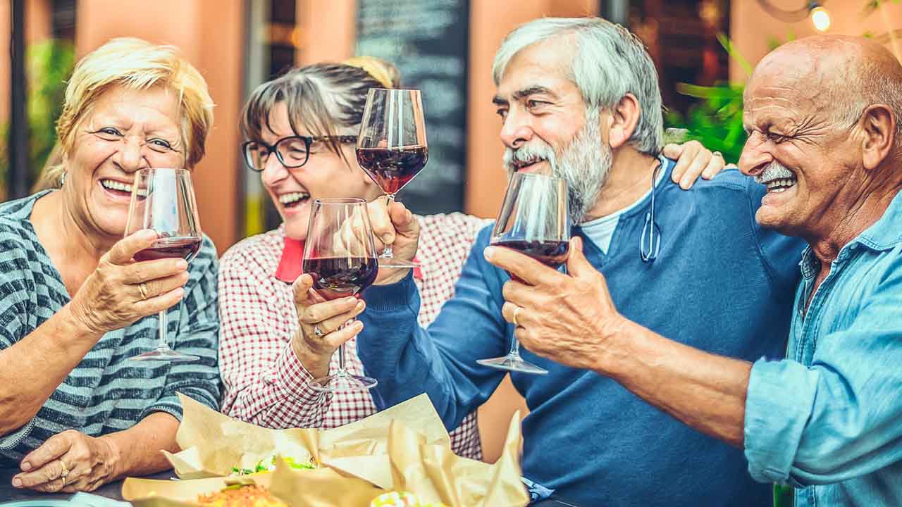 Four older adults sit together at a table outdoors, smiling and laughing as they toast with glasses of red wine. Plates of food are in front of them, creating a warm, joyful atmosphere.