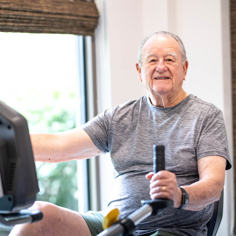 An older man wearing a gray t-shirt and shorts smiles while exercising on a rowing machine indoors, near a window with natural light.