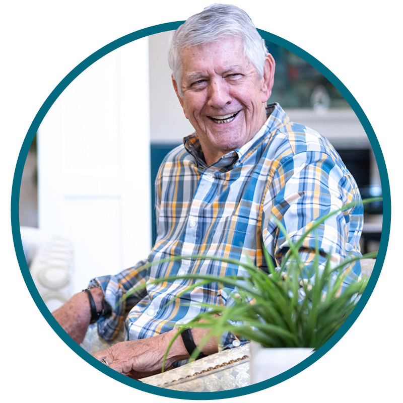 Smiling older man with short gray hair wearing a blue, yellow, and white plaid shirt sits indoors. There is a green potted plant in the foreground. The image is framed in a circular border.