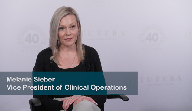 A woman with straight blonde hair sits in a chair, wearing a black top and light pants. A text overlay reads "Melanie Sieber, Vice President of Clinical Operations." The background features a faint logo and a "40" anniversary symbol.