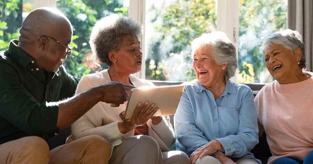 Four older adults sit together on a couch, smiling and laughing while looking at a tablet device, with sunlight streaming through large windows behind them.