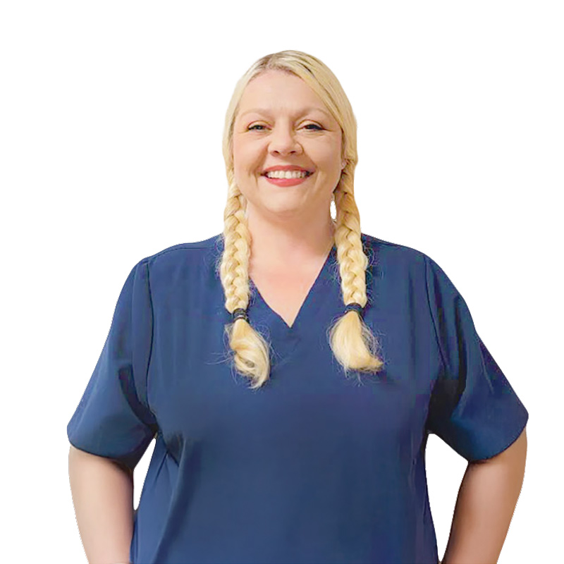 A woman with blonde hair in two braids, wearing a navy blue scrub top, stands smiling with her hands on her hips against a plain white background.