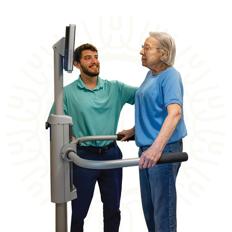 A man assists an older woman standing at a medical device with handrails and a screen, both smiling and focused on the equipment. The background is white with a subtle abstract pattern.
