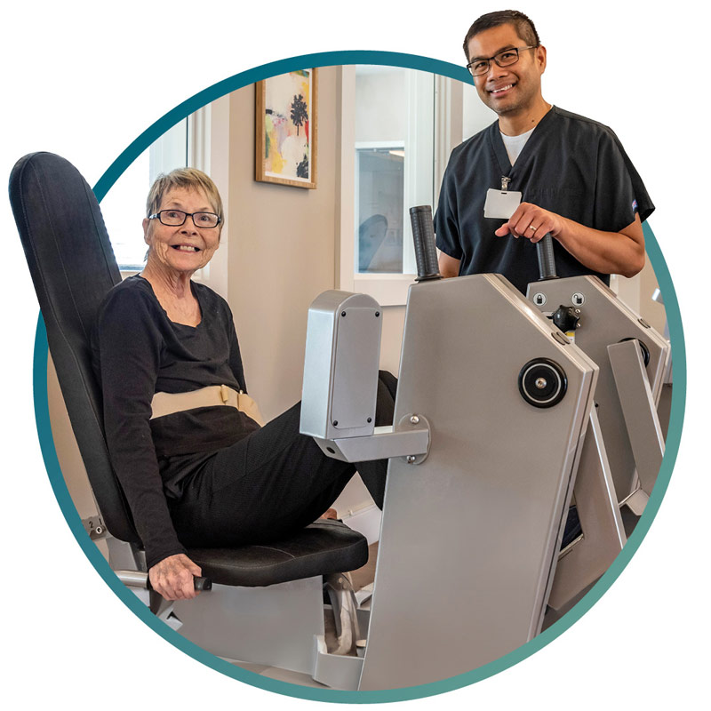 An older woman uses a leg press machine while smiling at the camera, assisted by a healthcare professional standing beside her in a clinic setting.