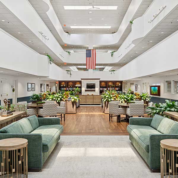 Spacious, modern lobby with green sofas, wooden floors, plants, a large American flag hanging from the ceiling, and abundant natural light from skylights above.