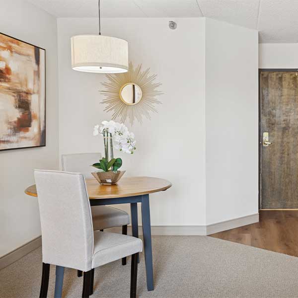 A small dining area with a round table, two upholstered chairs, a potted white orchid, wall art, a sunburst mirror, and a hanging light fixture near a wooden door.