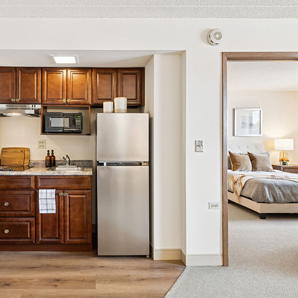 A small kitchen with wood cabinets, a silver fridge, microwave, and sink is next to an open doorway leading to a bedroom with a made bed, nightstand, and lamp.