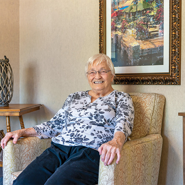 An elderly woman with short white hair and glasses smiles while sitting in a patterned armchair. A colorful framed painting hangs on the wall behind her.