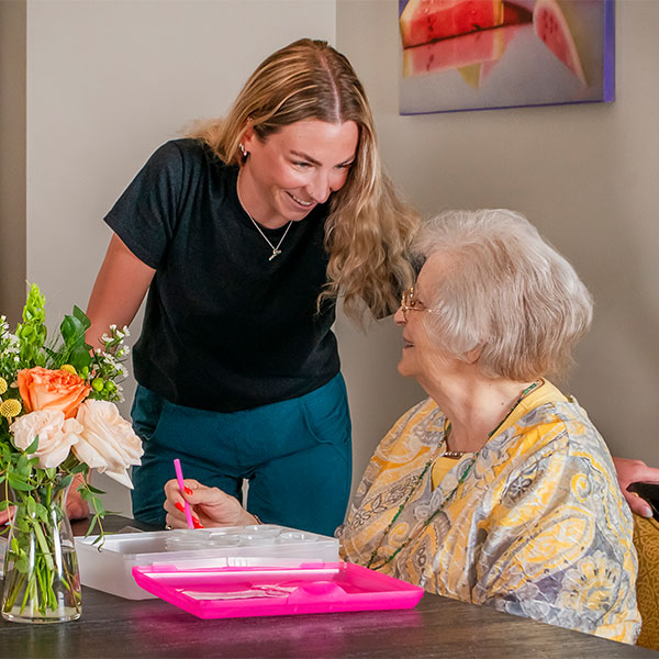 A younger woman smiles at an older woman who is seated and holding a pen. They are at a table with a bouquet of flowers and pink trays. A painting of watermelon hangs on the wall behind them.
