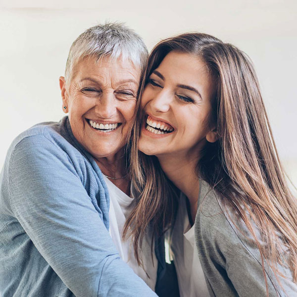 Two women, one older with short gray hair and one younger with long brown hair, smile and hug each other while looking at the camera, expressing happiness and warmth.