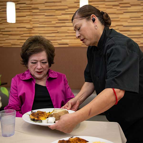 A woman in a pink jacket sits at a table as a server in black places a plate of food in front of her. A glass of water is on the table and the background features a patterned wooden wall.