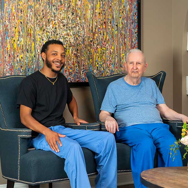 A young man and an older man sit side by side on blue chairs, both smiling. They wear blue pants and casual shirts. A colorful abstract painting hangs on the wall behind them.