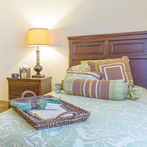 A neatly made bed with patterned pillows and a tray on top, next to a wooden nightstand with a lamp, small clock, and vase of white flowers. The room is softly lit and decorated in light, calming colors.