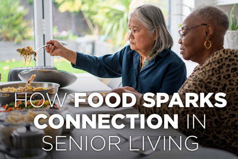 Two senior women sit at a table sharing food, serving noodles from a large bowl. Sunlight streams through a window behind them. Text reads, “How food sparks connection in senior living.” Kelly Hilmes Elected as community chairperson highlights the power of shared meals in bringing people together.