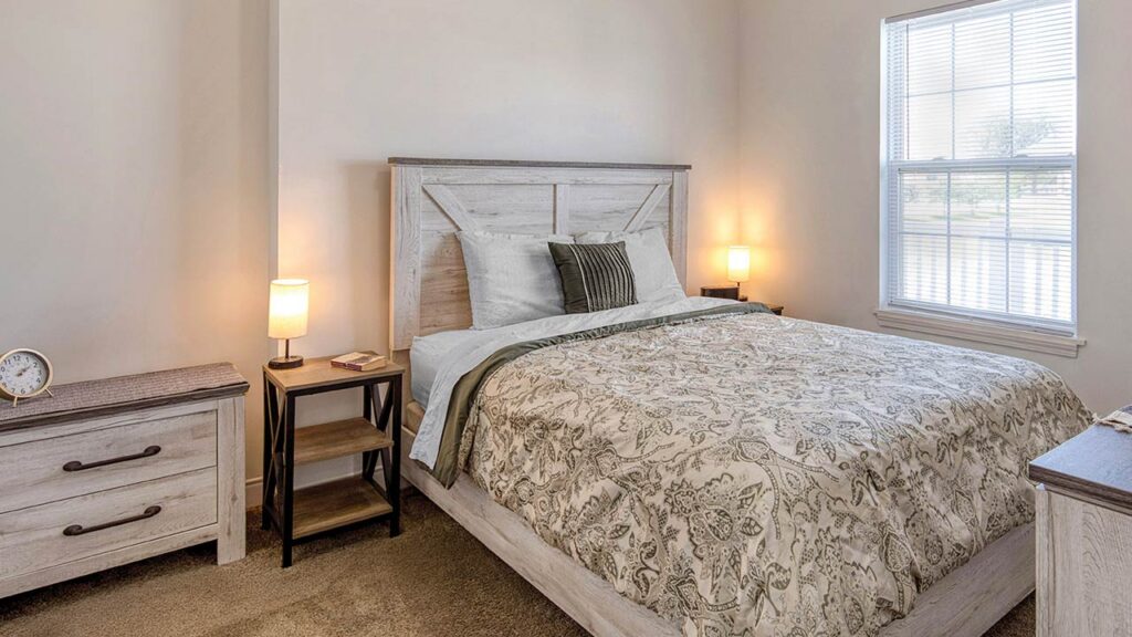 A cozy bedroom with a patterned bedspread, two lit lamps on nightstands, a chest of drawers, a clock, and a large window letting in natural light.