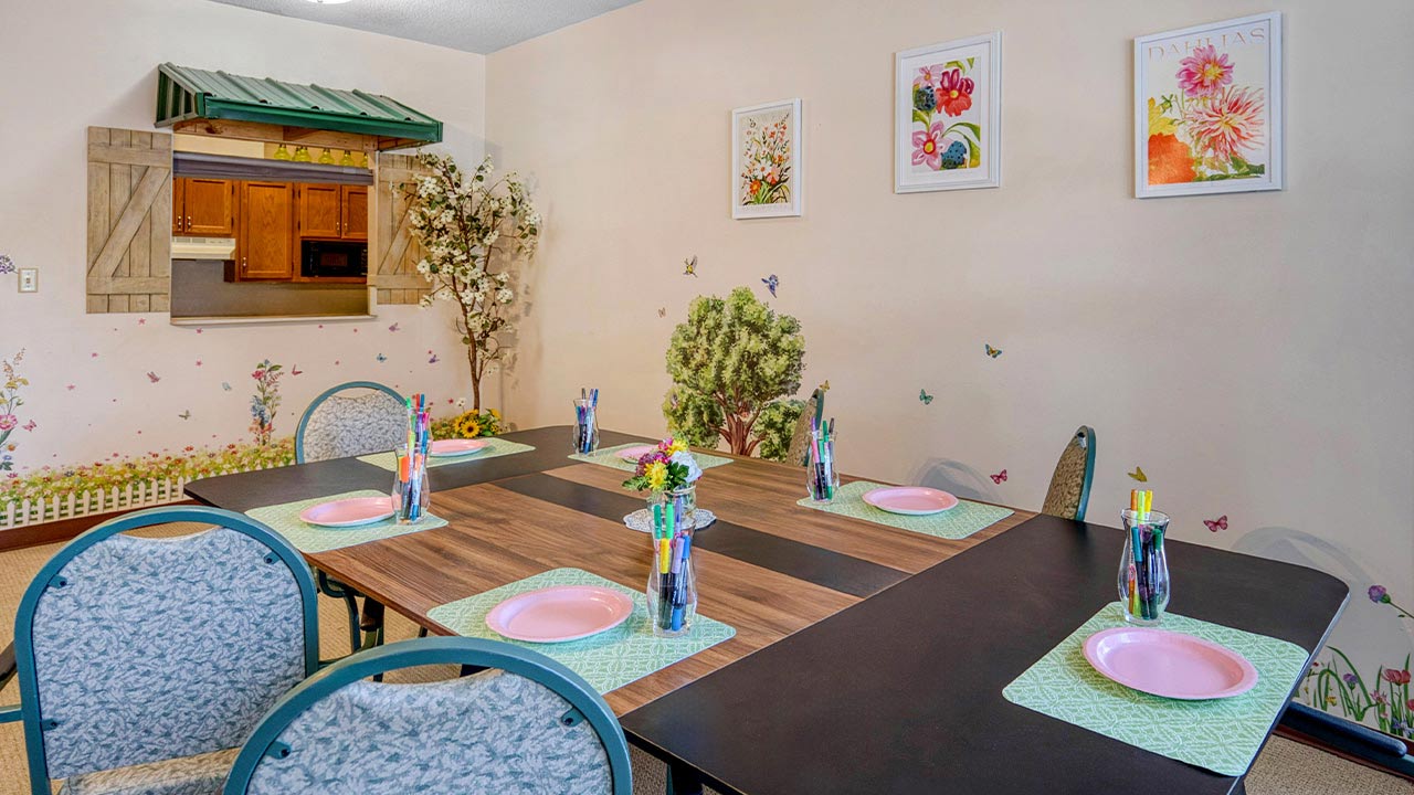 A brightly decorated room with a square table set for six, pink plates, green mats, and colorful cups. Art, faux flowers, and a window-like opening to a kitchen are visible. Butterflies and plants decorate the walls.