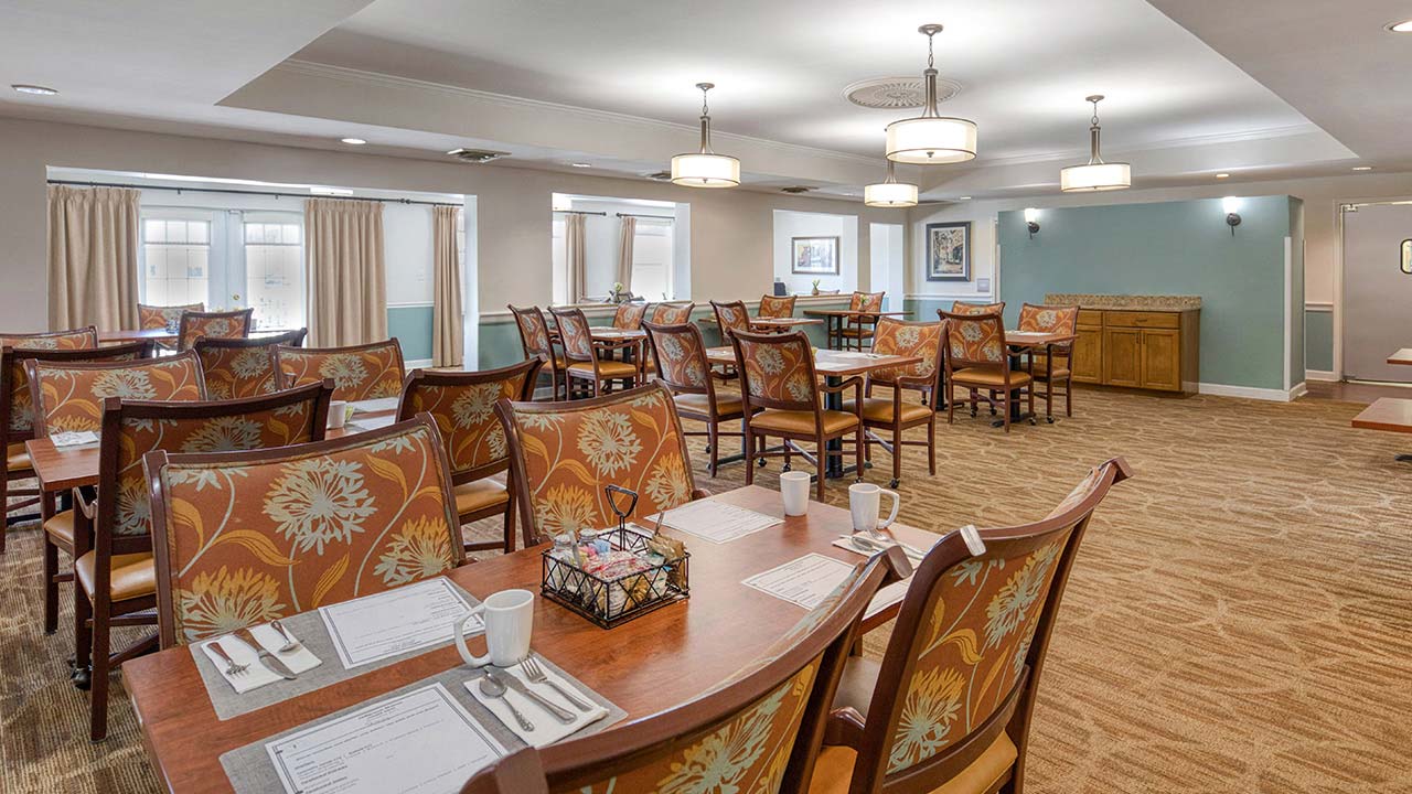 A spacious dining room with wooden tables and floral cushioned chairs, set with placemats, cups, and utensils. Large windows and ceiling lights brighten the room.