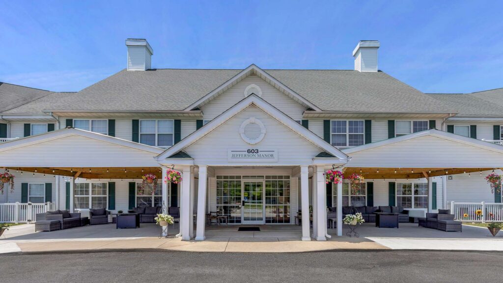 Two-story building with a covered entrance, large windows, outdoor seating, and hanging flower baskets. The sign above the door reads “603 Jefferson Manor.” The sky is clear and blue.