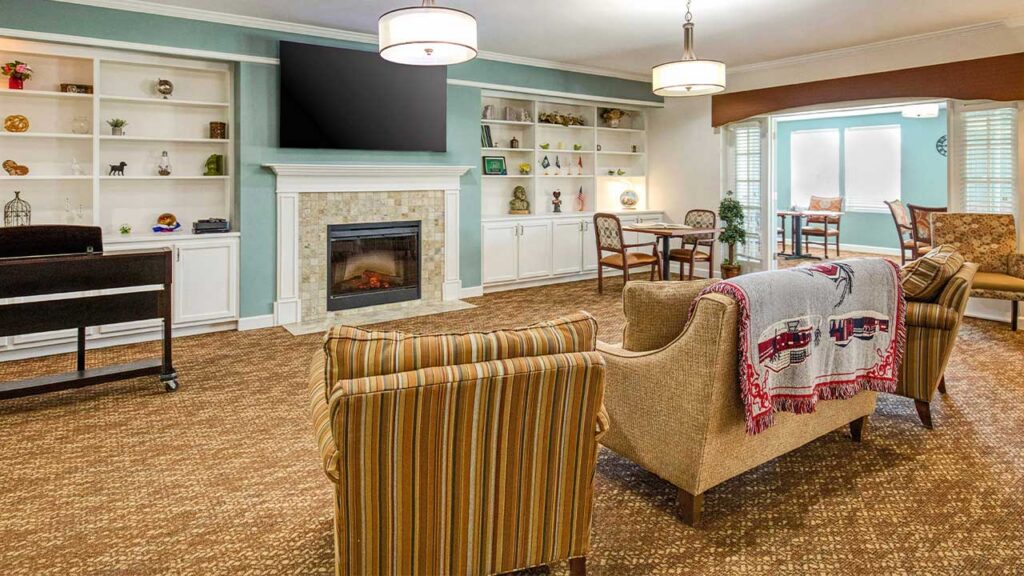 Cozy living room with striped chairs, a beige couch, wall-mounted TV, fireplace, built-in shelves with decor, and a dining table by the windows. The room is warmly lit and has brown carpeting.