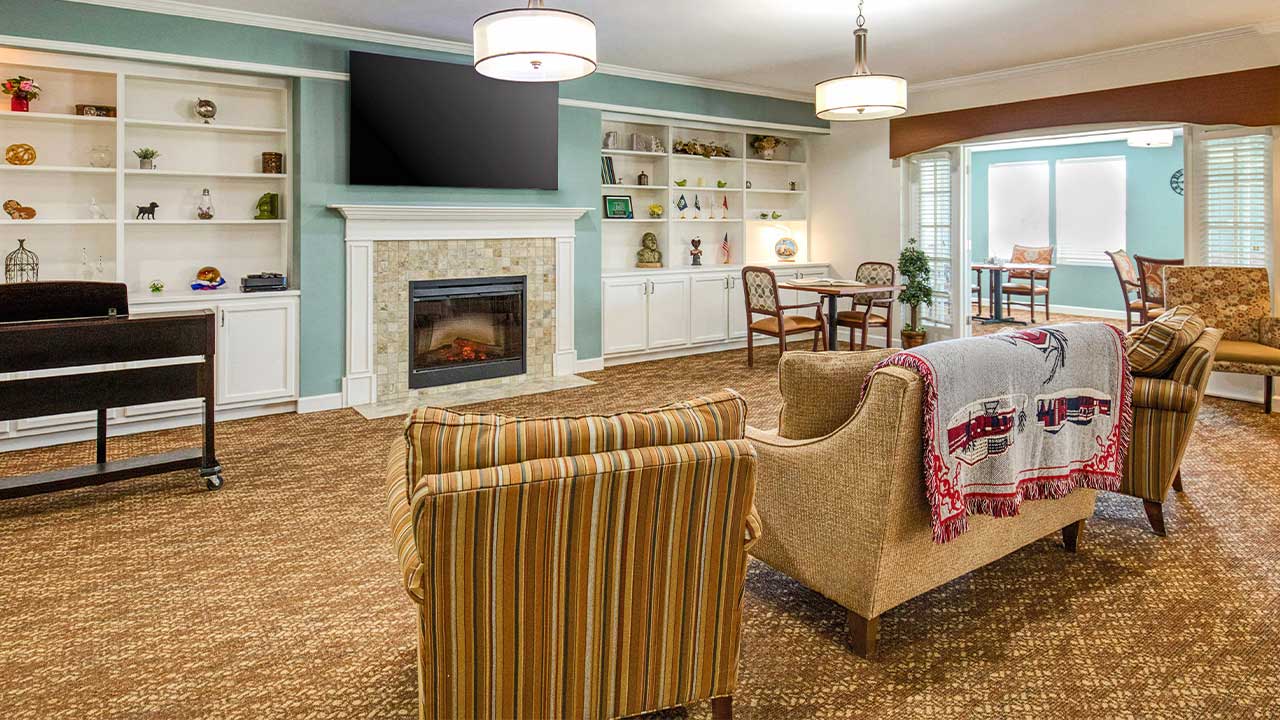 Cozy living room with striped chairs, a beige couch, wall-mounted TV, fireplace, built-in shelves with decor, and a dining table by the windows. The room is warmly lit and has brown carpeting.