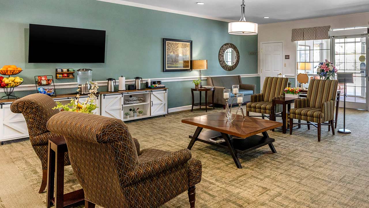 A cozy lounge with patterned armchairs, a coffee table, refreshments on a sideboard, a flat-screen TV, and a door leading to a sunlit area. Decorative items and greenery add warmth to the space.