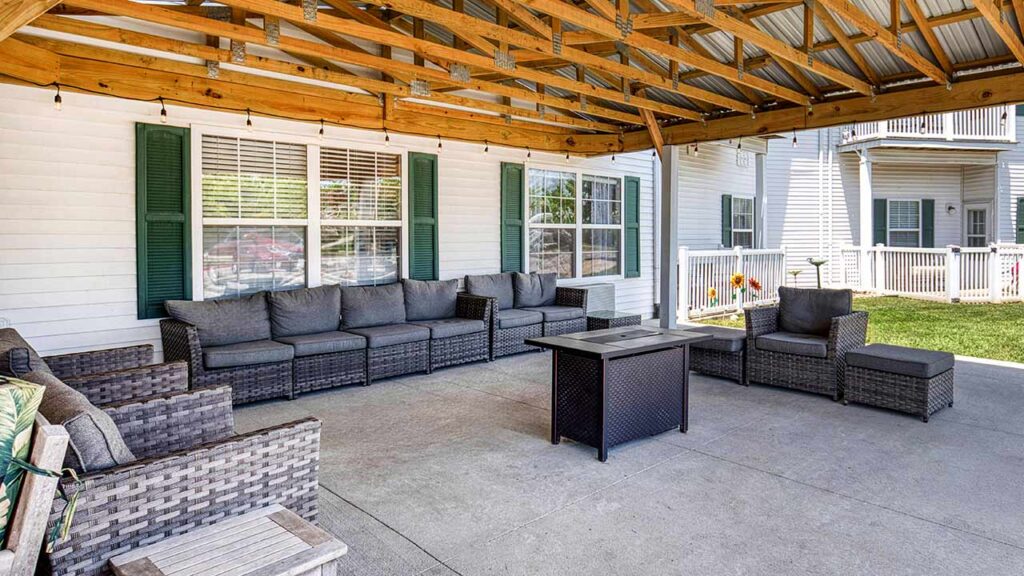 Outdoor covered patio with gray wicker sofas and armchairs, gray cushions, a rectangular fire pit table, string lights overhead, and a view of a house with green shutters and a yard.