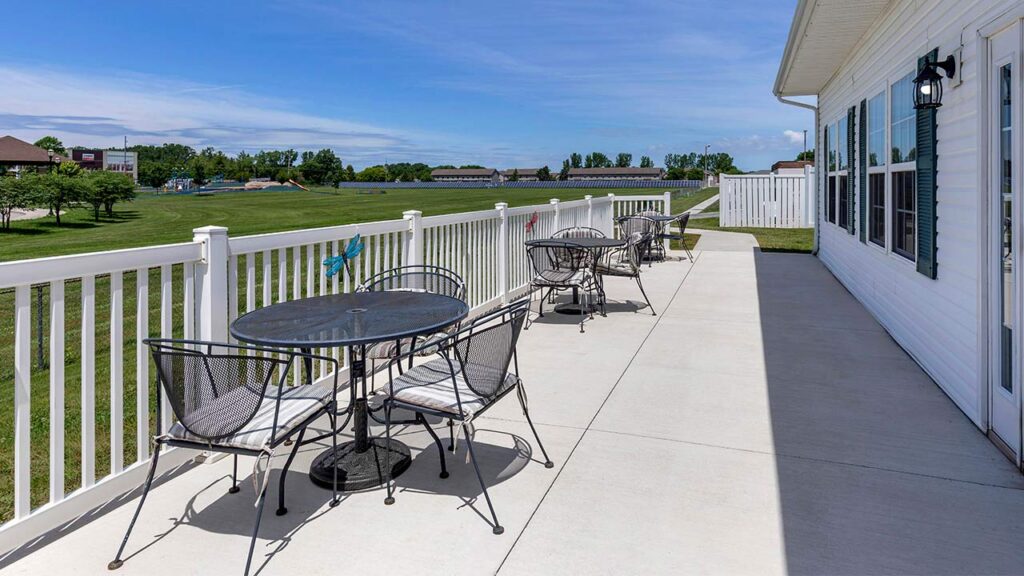 A long, white-fenced patio with metal tables and chairs runs alongside a white building. The patio overlooks a green lawn with trees and a pond under a blue sky.