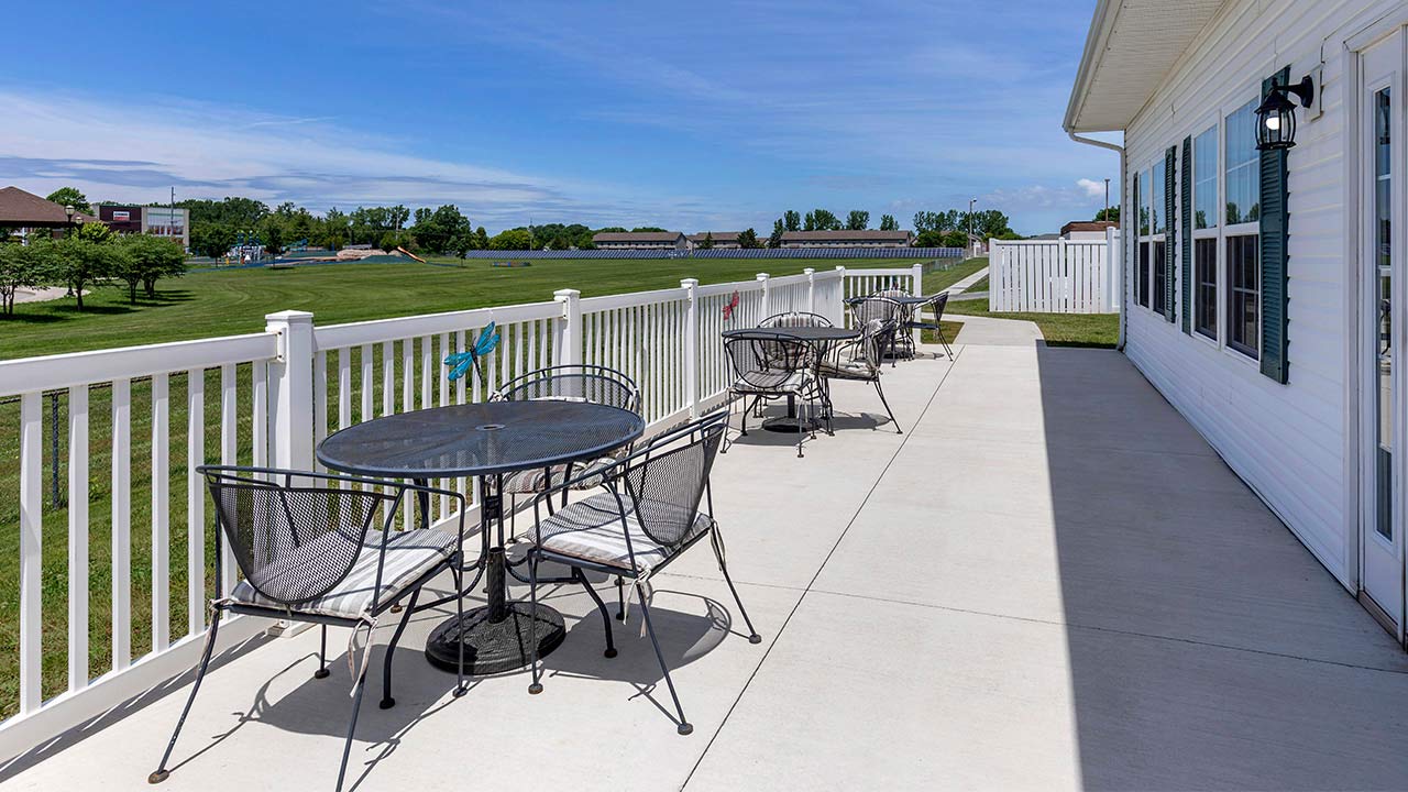 A long, white-fenced patio with metal tables and chairs runs alongside a white building. The patio overlooks a green lawn with trees and a pond under a blue sky.