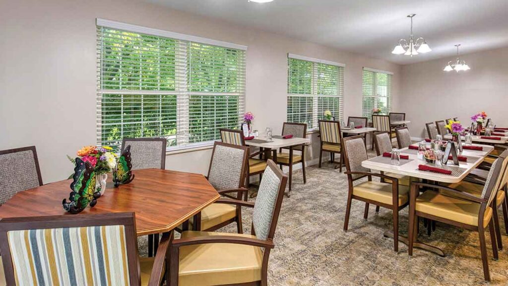 A dining room with multiple hexagonal and rectangular tables set for meals, surrounded by chairs and large windows with blinds.