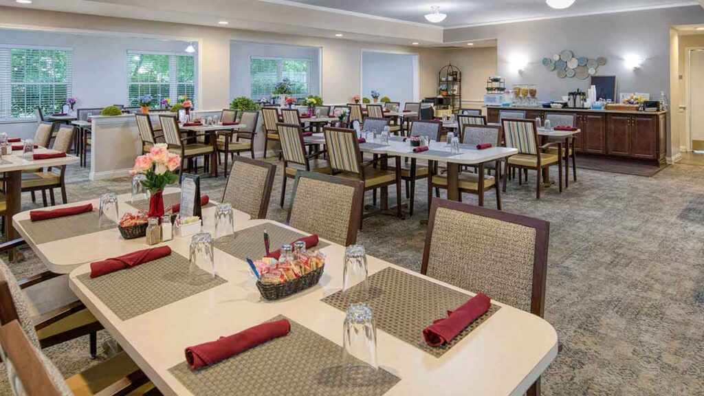 Dining room with multiple tables set for meals, each with place mats, napkins, and glasses, and a beverage station in the background.