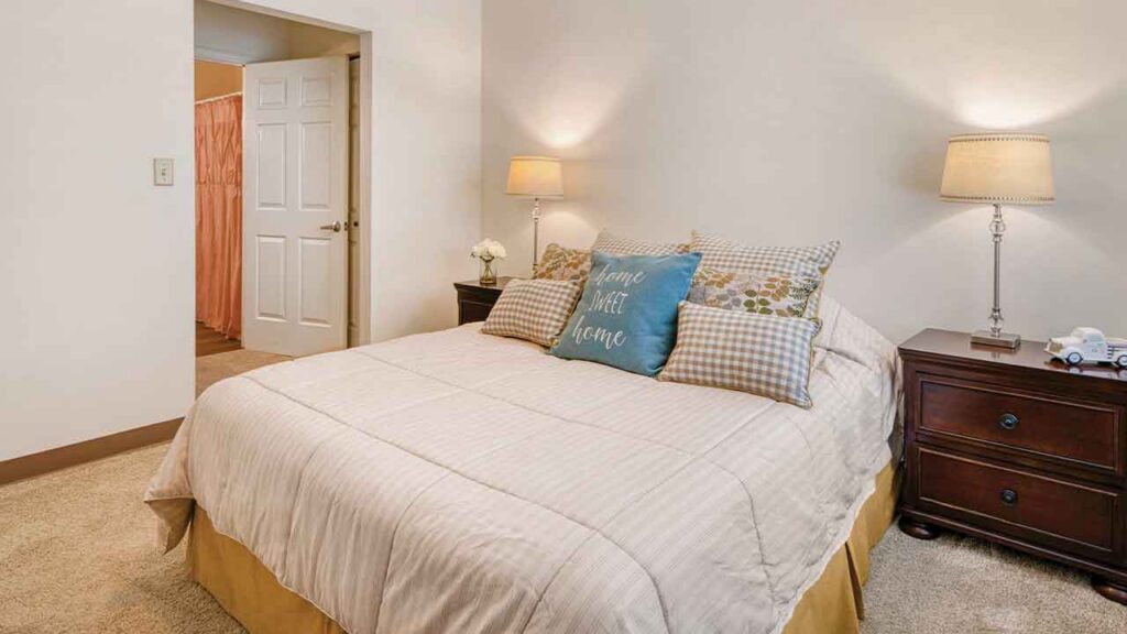 A queen bed with a light-colored quilt and several pillows, including one reading “home sweet home,” centered between two wooden nightstands with lamps.
