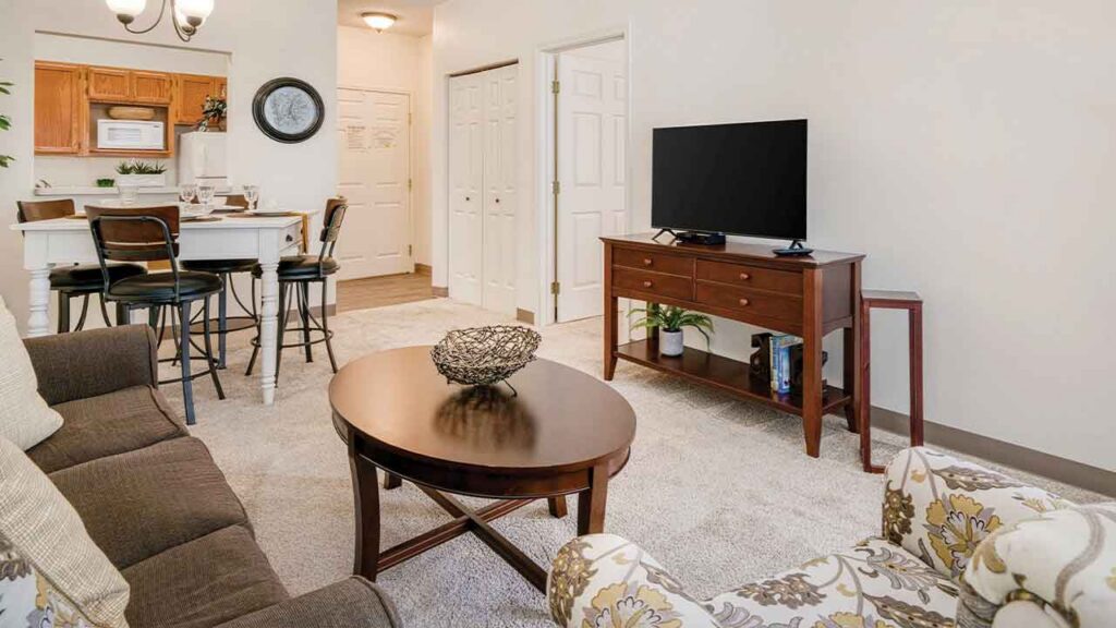 An open-concept living and dining area featuring a sofa, patterned armchair, coffee table, television on a wooden console, and a dining table with bar stools.