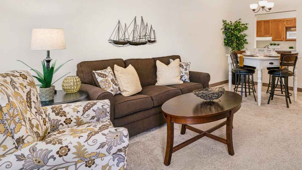 A living room with a brown sofa, a patterned armchair, an oval coffee table, and a dining area with a table and stools in the background.