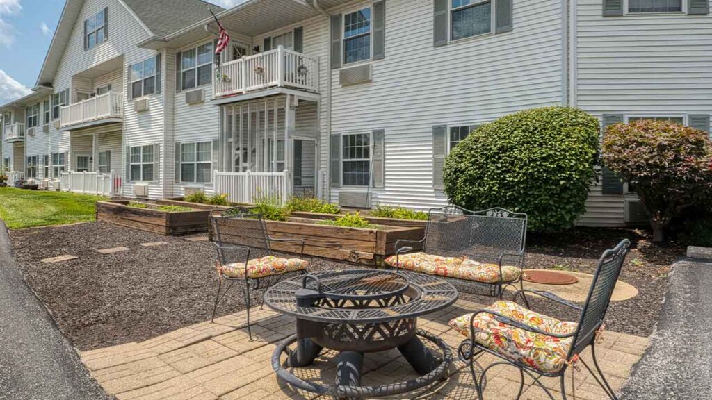 A patio with a fire pit, three chairs with patterned cushions, and two wooden raised garden beds in front of a white apartment building.