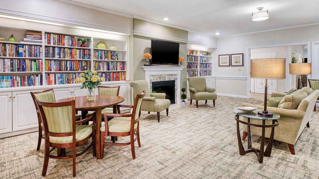 A common room featuring a dining table with chairs, two armchairs flanking a fireplace with a mounted television, a sofa, and floor-to-ceiling bookshelves.
